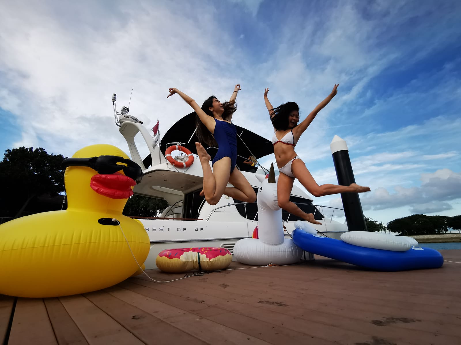 Two guests leaping between large inflatable floats on the deck beside a moored yacht, including an oversized rubber duck and a unicorn