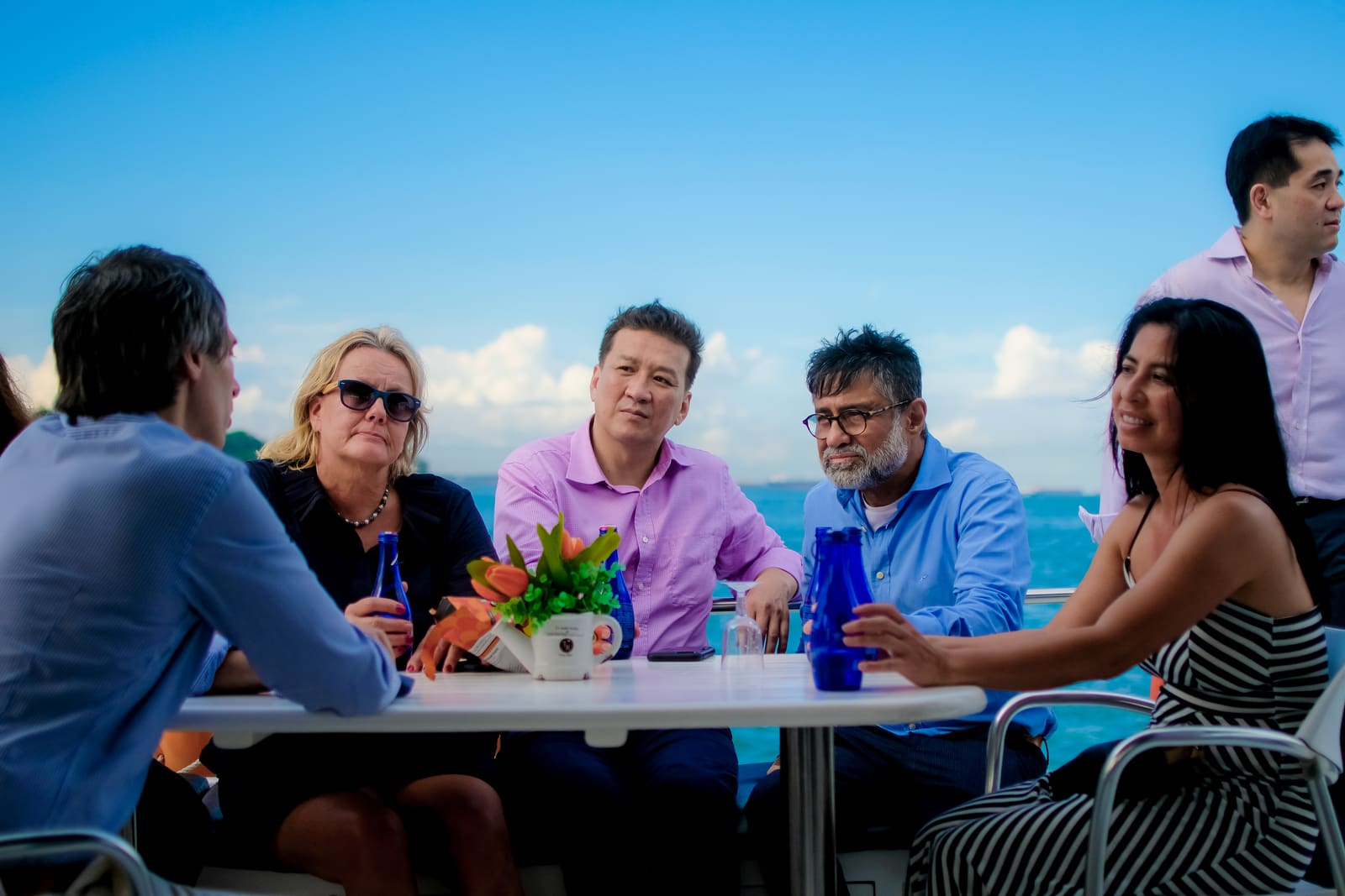 Seven colleagues seated around a table on the deck of a charter yacht in the late afternoon, drinks and a small plant on the table, open water behind them