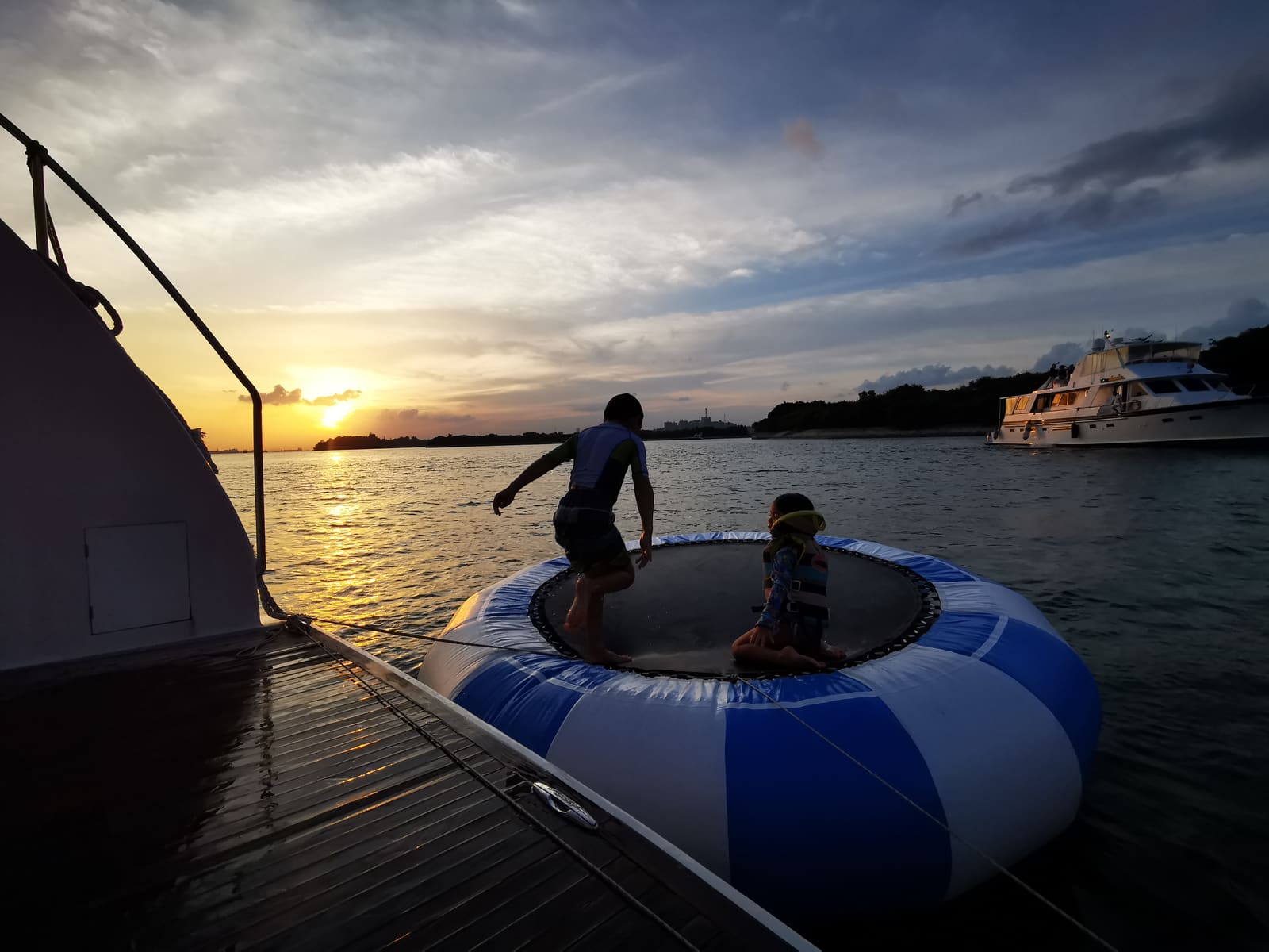Two children playing on an inflatable trampoline tethered off the stern at dusk, the younger one in a life jacket, with the sun setting behind an island