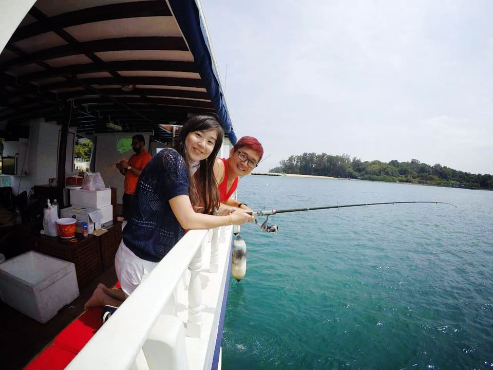 Two guests smiling over the white side rail of a charter boat with a spinning rod out over clear green water, a crew member rigging a line under the canopy behind them and a wooded island in the distance