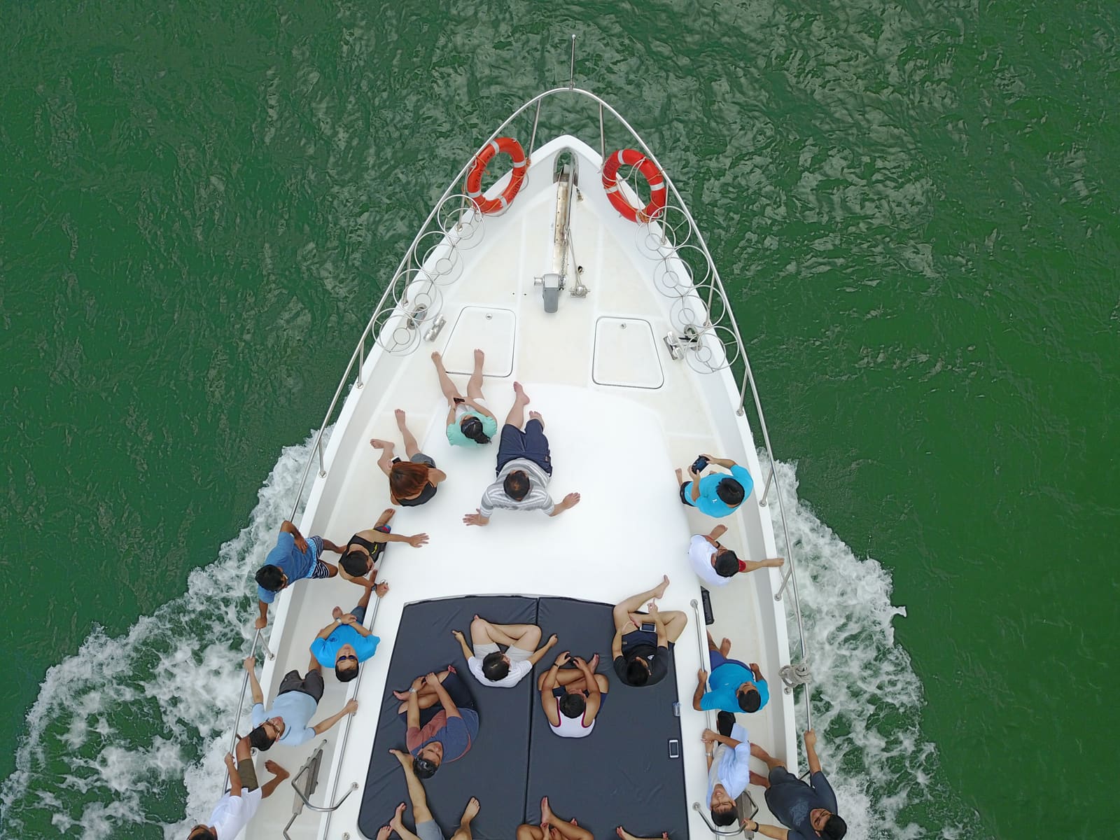 Overhead view looking down on the bow of a motor yacht under way, with around twenty guests seated across the foredeck and sun pad