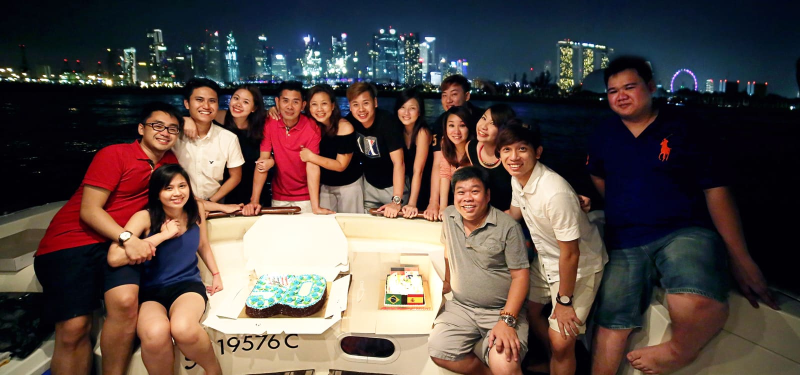 A group of about fifteen gathered around a birthday cake on the aft deck at night, with the lit city skyline and the Singapore Flyer behind them