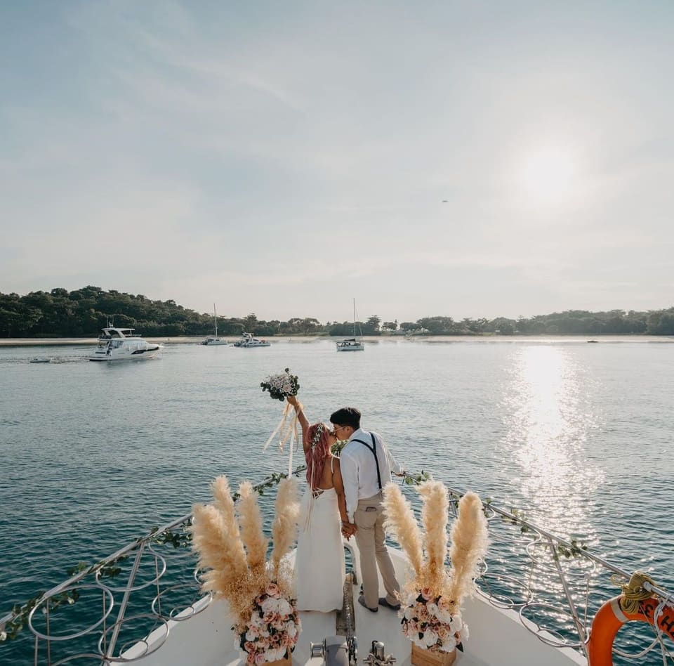 A couple embracing on the decorated bow of a yacht at anchor in late afternoon light, pampas grass and flowers along the rails, an island and moored boats behind