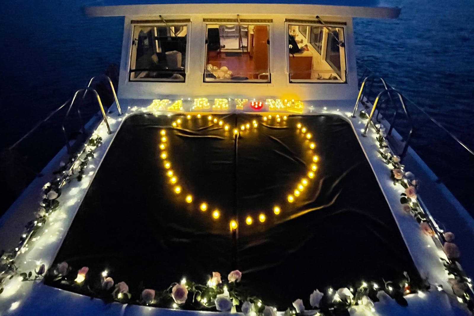 A yacht foredeck set up for a proposal after dark, with a heart of candles, Marry Me spelled out in lights, and flowers and fairy lights along the bow rails