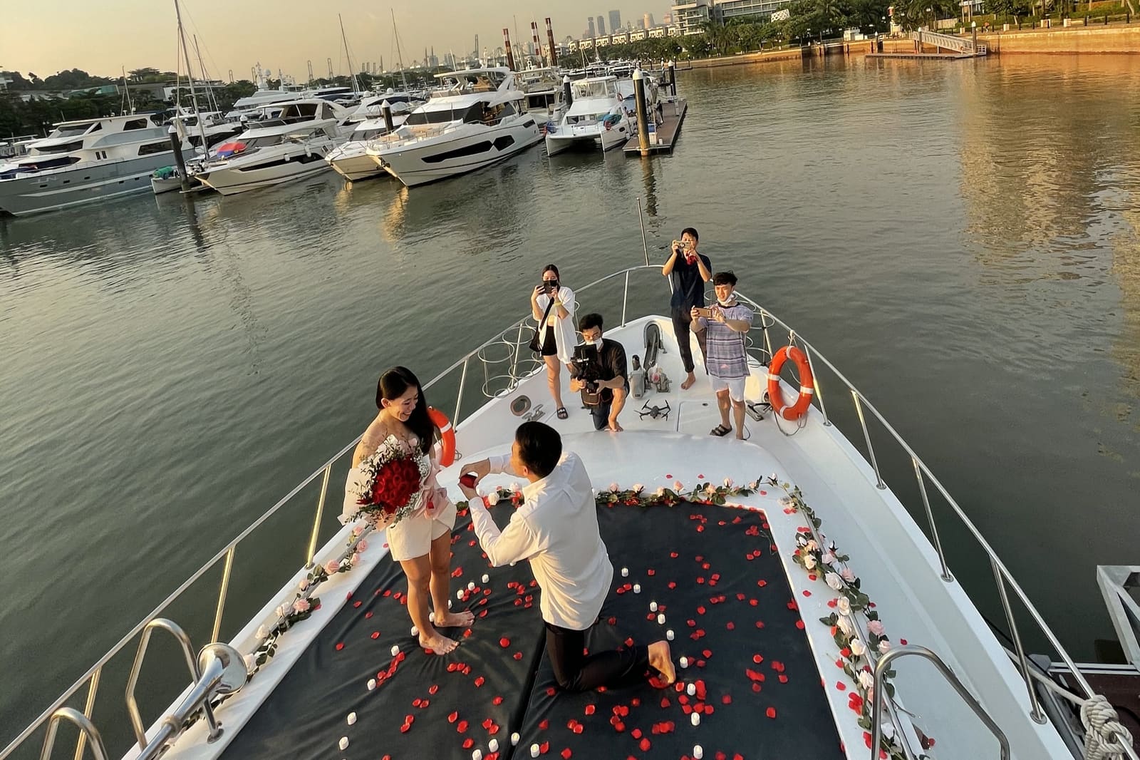 A man on one knee proposing on the foredeck of a yacht at golden hour, rose petals scattered across the deck, a photographer and friends looking on, marina behind