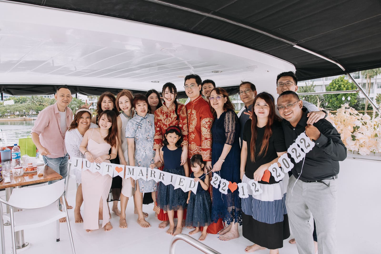 A wedding party of around eighteen gathered on a shaded aft deck, the couple in red and gold traditional dress, guests holding a Just Married banner