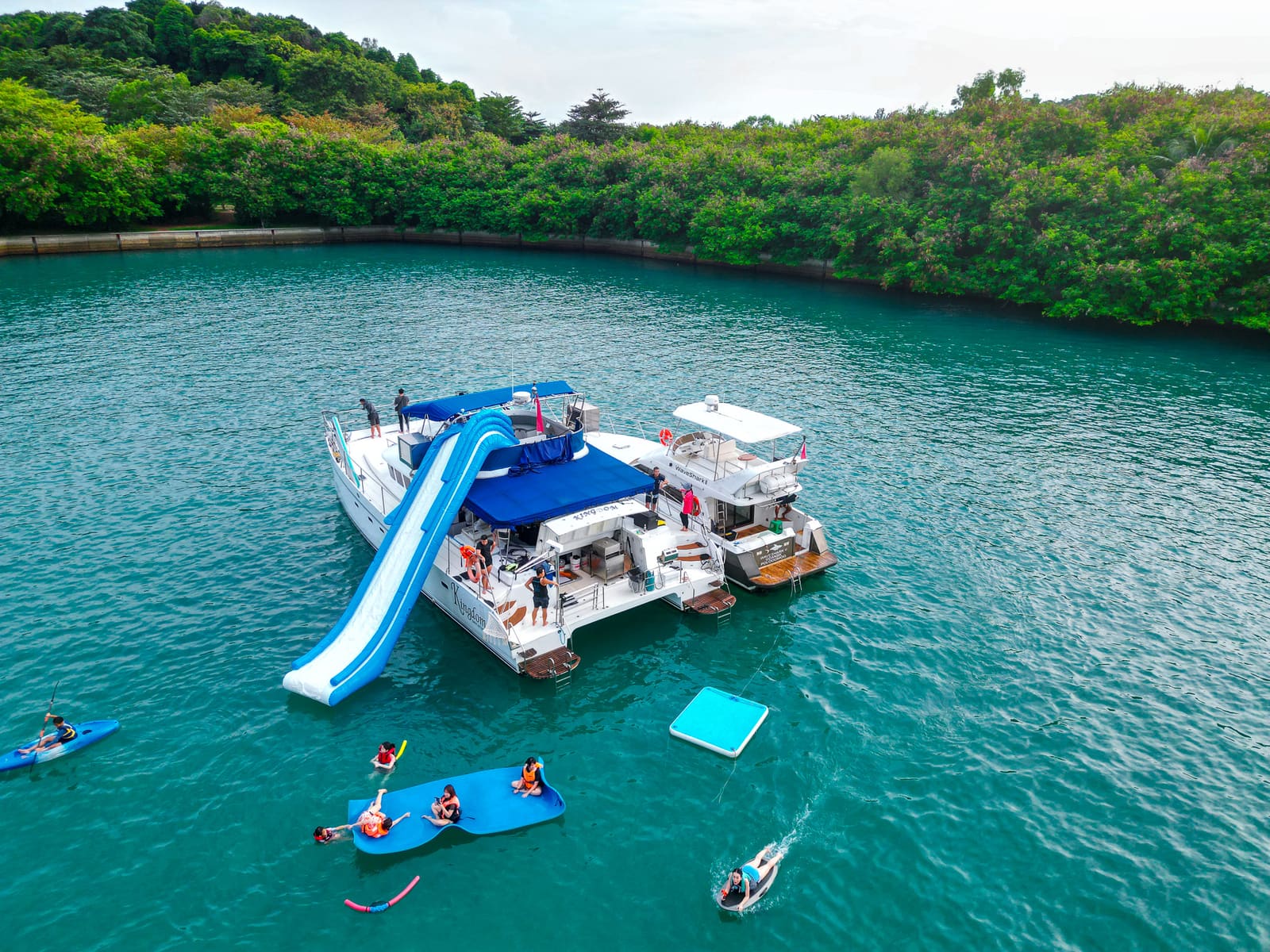 Aerial view at the anchorage — the slide down her starboard side, with kayaks, a paddleboard and the floating mat in the water