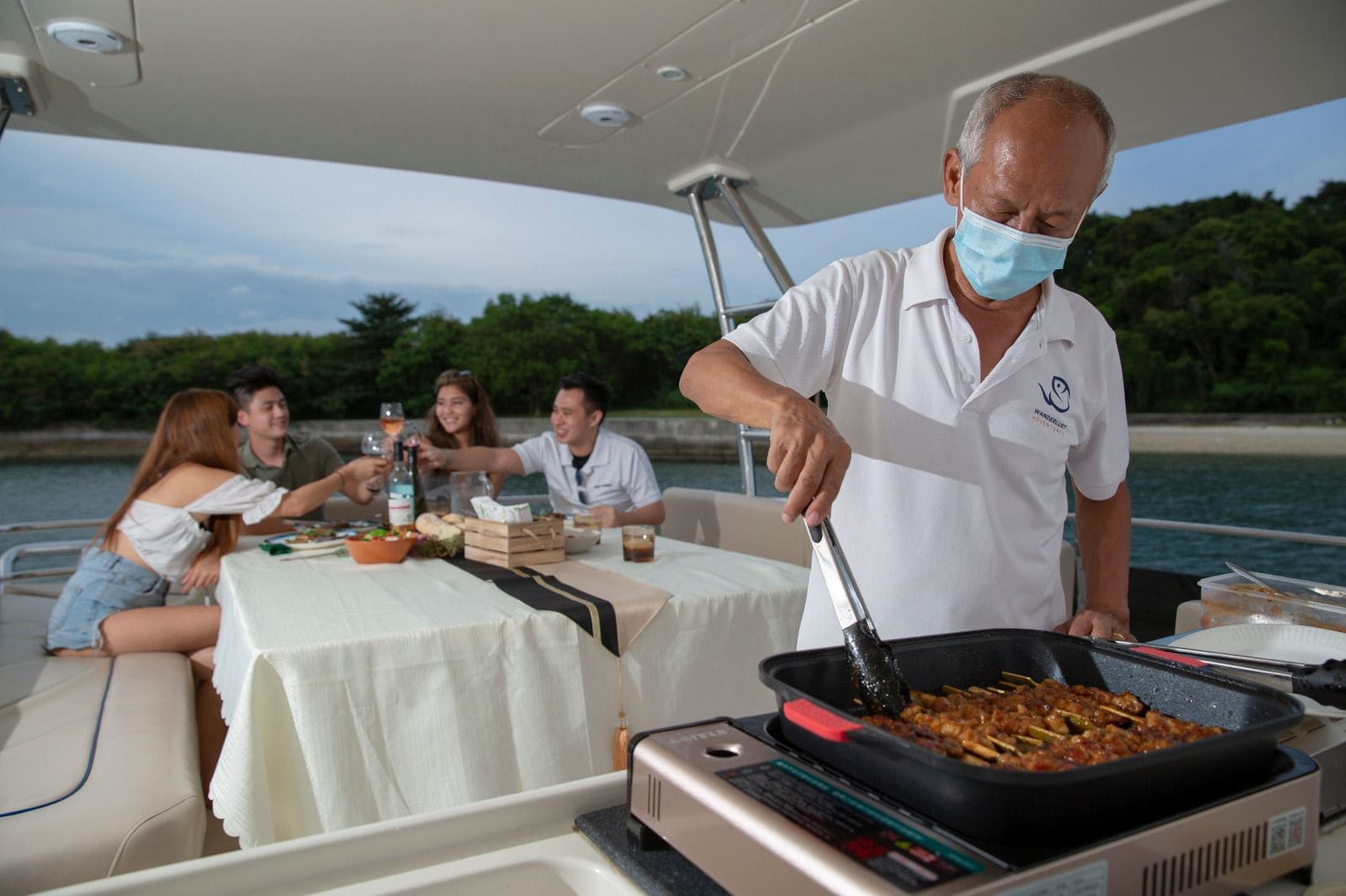 Crew grilling satay on the aft deck while guests eat at a laid table behind, the BBQ-with-service add-on in use