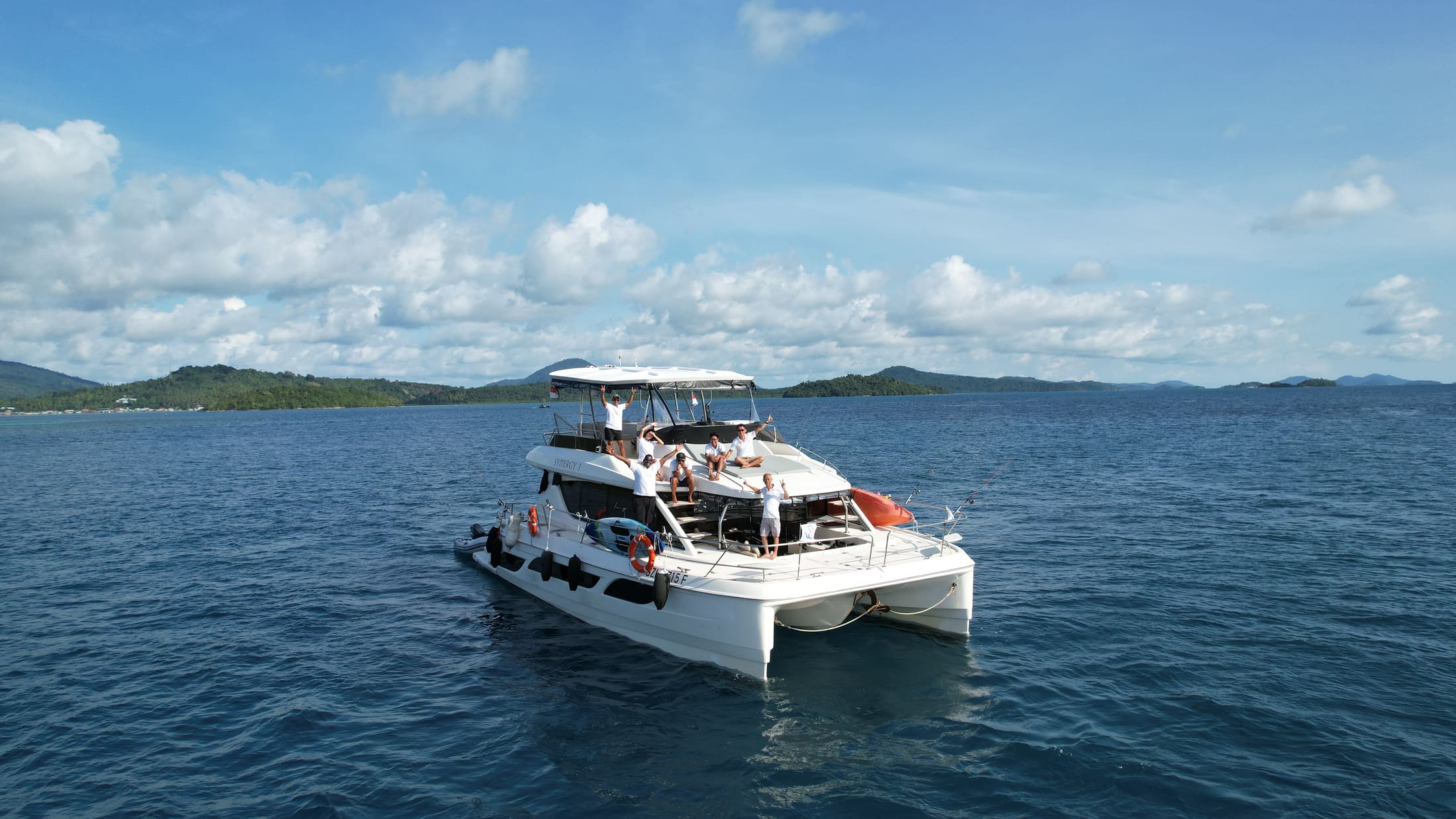 Synergy 1 from the air at anchor, guests waving from the flybridge and the foredeck, with both hulls and the hardtop clear against the water
